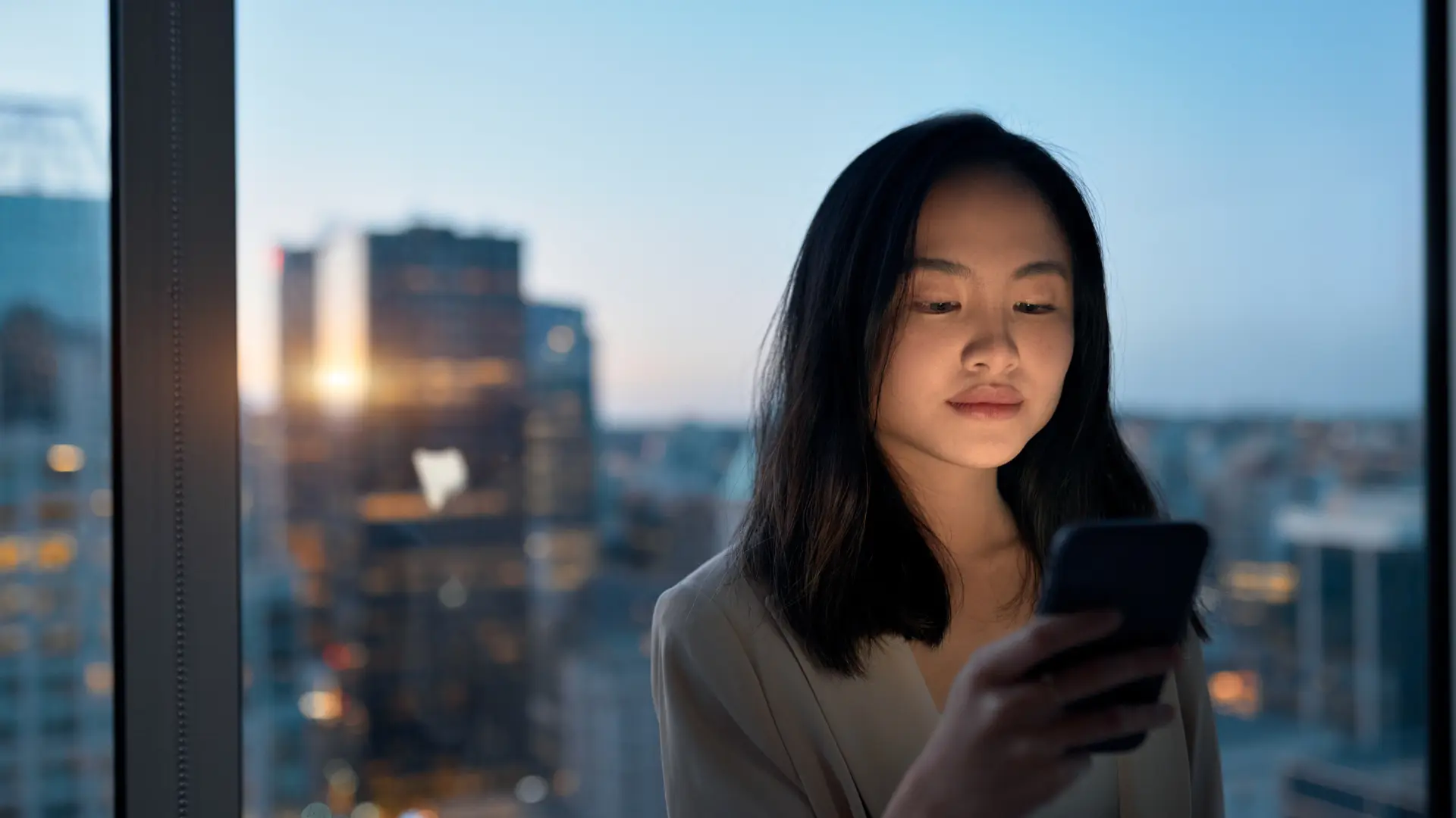 A young professional looking at her phone by a city window at dusk